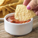 A person holding a chip and dipping it into a white Carlisle ramekin of salsa.