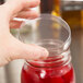A hand holding a clear Mason jar with a red liquid and an American Metalcraft Mason jar lid on the counter.