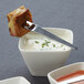 A white square porcelain sauce cup with a spoon in it on a table with a bowl of red soup.