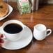 A CAC white bone china coffee cup on a wooden surface with a cup of coffee and a plate of bagel.
