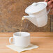 A white CAC Harmony teapot pouring liquid into a white cup.
