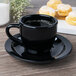 A black GET Elegance espresso cup and saucer on a wooden surface with small pastries.