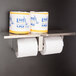 A Bobrick multi roll toilet paper dispenser with a metal shelf holding two rolls of toilet paper.
