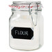 A glass jar of flour with an American Metalcraft rectangular chalkboard label on it.