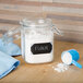 A glass jar of white powder with a rectangular chalkboard label next to a blue measuring spoon.