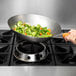 A hand using a Town mandarin carbon steel wok to cook carrots and broccoli in a pan on a stove.