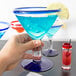 A hand holding a Libbey martini glass with blue liquid and a lemon wedge.
