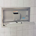 A Koala Kare grey baby changing station with a stainless steel flange on a white tile wall.