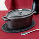 An American Metalcraft pre-seasoned black cast iron oval Dutch oven with a lid on a table with a red tablecloth.
