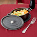 A pre-seasoned American Metalcraft mini cast iron oval Dutch oven with a lid filled with vegetables on a table with a fork.