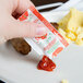 A hand holding a Ketchup packet over a plate of food.