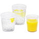 Three Dinex clear plastic tumblers filled with orange juice and garnished with lemon slices on a table with a bowl of lemons.