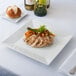 A 16" square white porcelain plate with chicken and vegetables on a table.