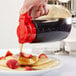 A hand holding a Tablecraft Dispenser Jar with a red top pouring syrup on a plate of pancakes.