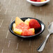 A Carlisle black rimmed melamine fruit bowl with a spoon on a table with a bowl of fruit.
