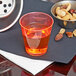 A Fineline Quenchers neon red plastic shot cup filled with orange liquid on a table next to a bowl of nuts.