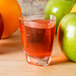 A triangle-shaped clear plastic GET dessert shot glass filled with orange liquid next to green apples.