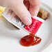 A hand holding a small Americana Ketchup packet over a plate.
