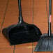 A black Rubbermaid Lobby Pro dustpan on a tile floor next to a broom.