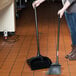 A woman using a Rubbermaid Lobby Pro dustpan and broom on a tile floor.