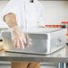 A person in gloves using a Vollrath aluminum roasting pan to put food on a table.