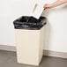A person's hand putting a white rectangular object into a beige Continental 25 gallon trash can with a black lid.