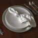 A Tabletop Classics silver plastic charger plate on a table with silverware and a napkin.