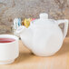 A white Tuxton teapot with a lid next to a white cup of tea on a wooden table.