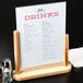 An American Metalcraft natural wood-finish table top board holding a menu on a stand on a table with a glass of liquid.