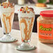 A jar of REESE'S Peanut Butter Sauce on a table next to two glasses of peanut butter milkshakes.