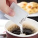 A person pouring sugar into a cup of coffee using a white packet from a Double Serving Hot Beverage Condiment Kit.
