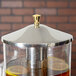 A Vollrath brass knob on a glass beverage dispenser filled with brown liquid and lemon slices.
