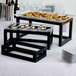 A group of black wood open frame risers holding trays of food on a table.