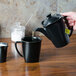 A person pouring liquid from a black teapot into a Hall China black mug.