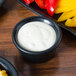 A black Hall China ramekin filled with white sauce next to a plate of red and yellow bell peppers.