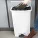 A woman using a white Rubbermaid Slim Jim trash can with a white lid to put a black box inside.