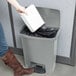 A woman using her foot to open a Rubbermaid Slim Jim trash can to put a white box inside.