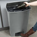 A woman using a black shoe to open a Rubbermaid Slim Jim front step-on trash can.