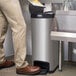 A person standing next to a Rubbermaid Slim Jim stainless steel end step-on trash can.