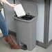 A woman using the front step pedal to open a Rubbermaid Slim Jim trash can to put a book inside.