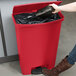 A woman using a Rubbermaid Slim Jim red front step-on trash can to throw away a black container.