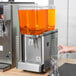 A person pouring orange liquid into a Crathco refrigerated beverage dispenser.