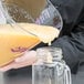 A person using a Cambro purple polycarbonate measuring cup to pour liquid into a glass jar.
