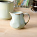 A Tuxton TuxTrendz sagebrush green china creamer on a table next to a cup of coffee.