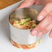 A hand holding a stainless steel round food mold filled with food.
