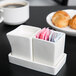 A white Tuxton china ramekin tray with sugar packets in it on a counter next to a plate of croissants.
