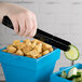 A hand using Fineline black plastic serving tongs to pick up a cucumber slice over a blue container of croutons.