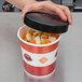 A hand holding a Choice paper soup container filled with food and covered with a black lid.