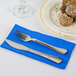 A fork and knife on a Creative Converting cobalt blue paper dinner napkin next to a plate of food.