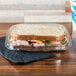A sandwich in a clear plastic Tamper Evident Take Out Container on a table in a deli.